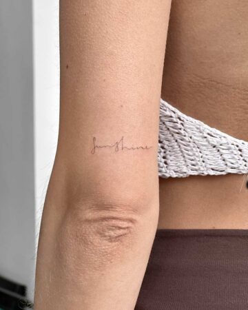 Delicate "Sunshine" script tattoo on a woman's arm, near the elbow. The fine-line tattoo is in a simple, elegant font, showcasing a minimalist tattoo design. This sunshine tattoo is a beautiful example of script tattoos. done by artists in our fine line tattoo studio Canguu Bali