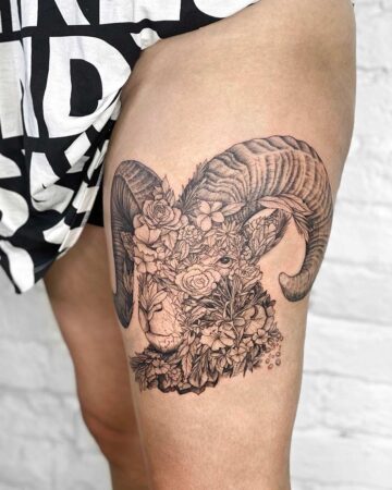 Detailed ram tattoo on a thigh, featuring intricate floral elements and curved horns. The black ink design showcases a ram's head surrounded by roses, lilies, and other flowers, creating a unique floral tattoo. This body art is a stunning example of a thigh tattoo. Done by tattoo artists in our fine line tattoo studio parlour shop Canggu, Bali