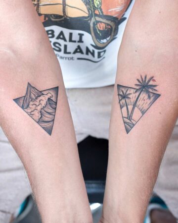 Two matching triangle tattoos on forearms, one depicting a wave crashing against a mountain, the other featuring palm trees and a sunset. Perfect best friend tattoos or couple tattoos. "Bali Island" shirt in the background. Done by tattoo artists in our fine line tattoo studio parlour shop Canggu, Bali