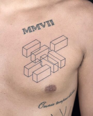 A man's chest features a geometric tattoo composed of interlocking 3D block shapes, inked in black. Above the design, the roman numerals "MMVII" are tattooed. Below, the phrase "Omnia tempus habet" is visible. A unique chest tattoo design. Done by tattoo artists in our fine line tattoo studio parlour shop Canggu, Bali