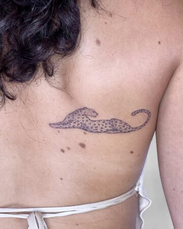 A minimalist leopard tattoo, featuring a reclining pose, is inked on a woman's back. The tattoo showcases a delicate design with fine lines and small spots. The woman's skin tone is light, and the tattoo is placed near her shoulder blade. Done by tattoo artists in our fine line tattoo studio parlour shop Canggu, Bali