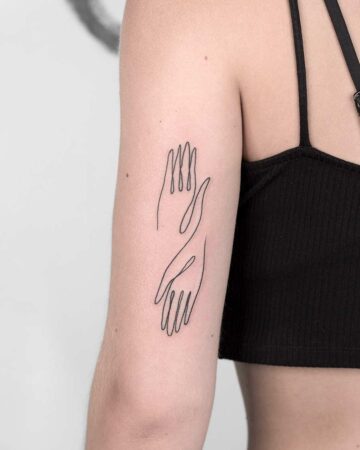 Minimalist line art tattoo of two hands reaching towards each other on a woman's upper arm. The black ink design features delicate lines, illustrating a connection or embrace. Done by tattoo artists in our fine line tattoo studio parlour shop Canggu, Bali