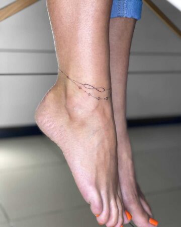 Delicate ankle tattoo featuring a minimalist infinity symbol and small circles, inked with fine lines. The tattoo sits on a woman's tanned ankle, with a second foot visible, and bright orange nail polish. Done by tattoo artists in our fine line tattoo studio parlour shop Canggu, Bali