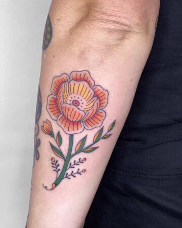 Vibrant orange and yellow floral tattoo on a forearm, featuring a large bloom with detailed petals, a green stem, and small leaves. The colorful tattoo is a beautiful example of a flower tattoo. Done by tattoo artists in our fine line tattoo studio parlour shop Canggu, Bali