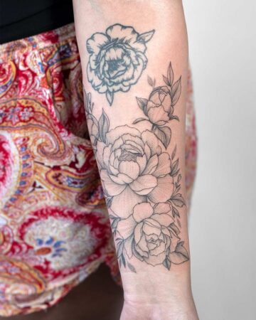 Detailed black and grey peony tattoo on a forearm, featuring three open blooms and several leaves, with a faded outline of a previous tattoo above. A beautiful floral tattoo example. Done by tattoo artists in our fine line tattoo studio parlour shop Canggu, Bali