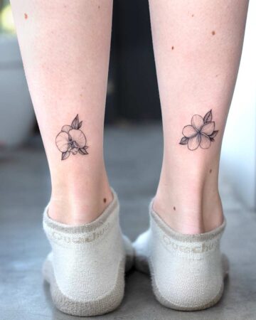 Two delicate flower tattoos grace the ankles of a person. The tattoos feature intricate line work, with one appearing to be an orchid and frangipany plumeria design and the other a cluster of petals. These minimalist flower tattoos are a beautiful example of small tattoo art.