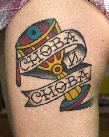 A colorful traditional tattoo on skin, featuring a banner with the Russian word "Snova" (again) in bold lettering. The tattoo has a detailed design with red, yellow, and blue elements, reflecting the style of traditional tattoo art. Done by tattoo artists in our fine line tattoo studio parlour shop Canggu, Bali
