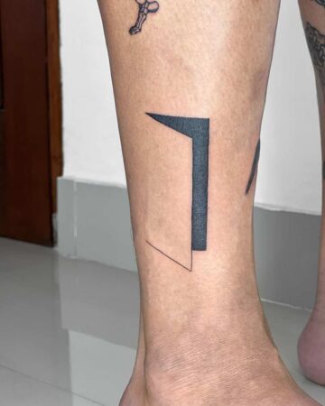 Minimalist geometric tattoo design on an ankle. A black ink tattoo features a stylized open door shape, with a solid black rectangle and triangle, and a thin black line. Done by tattoo artists in our fine line tattoo studio parlour shop Canggu, Bali