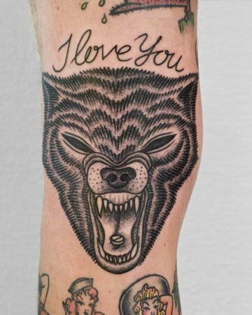 A detailed black ink wolf tattoo on an arm, snarling with bared teeth. Above the wolf's head, the words "I love you" are scripted in elegant cursive. The tattoo is a striking example of a wolf tattoo design. Done by tattoo artists in our fine line tattoo studio parlour shop Canggu, Bali