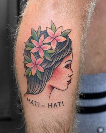 A colorful traditional tattoo of a woman's profile on a leg, adorned with flower blooms in her hair. The tattoo features black outlines, pink and green flowers, and the words "Hati - Hati" below.