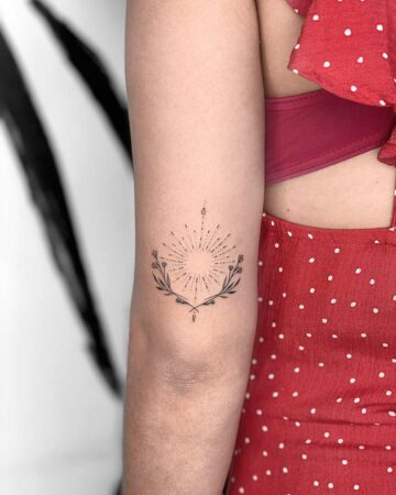 A delicate minimalist tattoo of a sunburst design framed by a floral wreath on a woman's arm. The small tattoo is inked in black, with fine lines and dots. The woman wears a red polka dot dress.