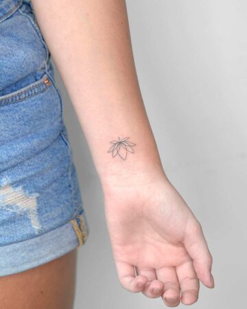 A minimalist lotus flower tattoo on a woman's wrist. The delicate black outline features open petals and a small dot detail, set against pale skin, with denim shorts in the background.