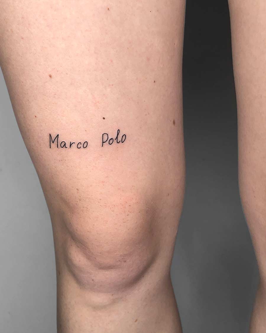 A minimalist script tattoo on a person's thigh reads "Marco Polo" in delicate, handwritten-style lettering. The tattoo is in black ink and is a small, subtle design.