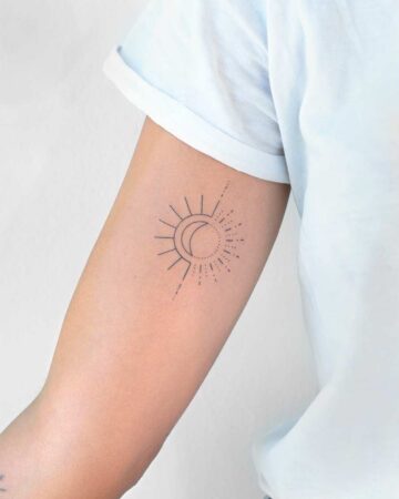 A minimalist sun and moon tattoo on a person's arm. The design features a sun with radiating lines and a crescent moon, connected by a dotted line. This small, fine line tattoo is a popular minimalist tattoo choice.