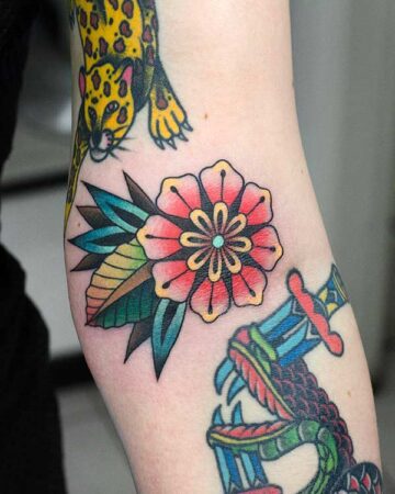 A vibrant, colorful flower tattoo adorns an arm, featuring red petals, yellow and black details, and blue-green leaves. A traditional tattoo style with a jaguar and snake/dagger tattoo visible nearby.