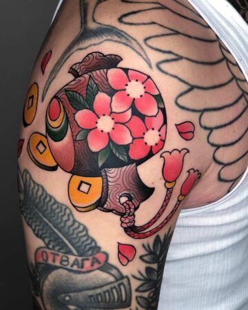 Bold tattoo of a Balinese Barong mask with a protruding tongue, surrounded by vibrant red and pink cherry blossoms on a man's upper arm. Japanese (Irezumi/Wabori) tattoo studio Canggu Bali