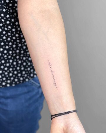 Delicate "tenderness" word tattoo in elegant script on a forearm, a minimalist design. Black bracelet on the wrist. Done by tattoo artists in our fine line tattoo studio parlour shop Canggu, Bali