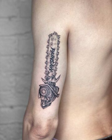 Detailed black and grey Chainsaw tattoo on an arm, featuring the word "PAINSAW" on the blade. The body art is intricately designed, with a mechanical aesthetic. Done by tattoo artists in our fine line tattoo studio parlour shop Canggu, Bali