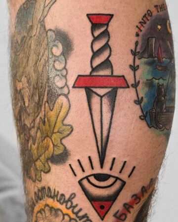 Detailed traditional tattoo art on a leg shows a dagger pointing down at an eye within a triangle. Red accents highlight the dagger's handle and the triangle's border. Other colorful tattoos surround the central image.
