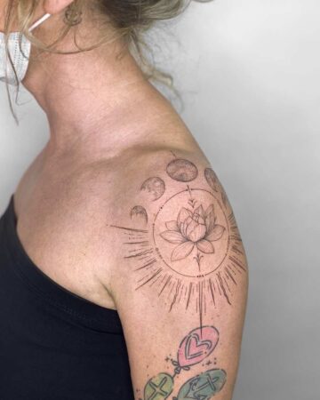 Shoulder tattoo featuring a delicate lotus flower surrounded by a circular design with moon phases and radiating lines, on a woman shoulder