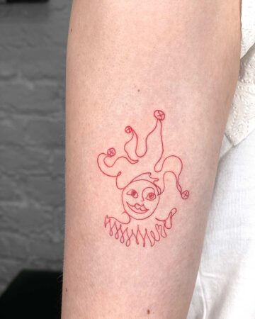 A minimalist fine line tattoo of a smiling jester in red ink adorns a person's arm. The design features a whimsical face, ruffled collar, and a playful, curly hat with small bells. This red ink tattoo is an example of fine line tattoo art. Done by tattoo artists in our fine line tattoo studio parlour shop Canggu, Bali