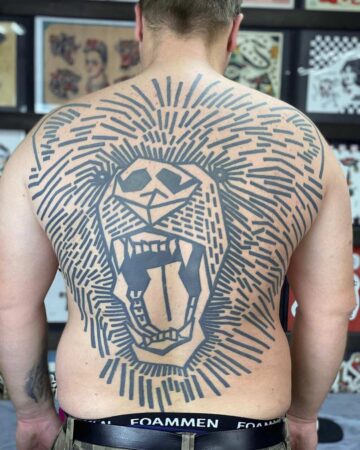 A man's back is covered with a large, geometric bear tattoo. The black ink design features a roaring lion's face with a sunburst-like mane, showcasing a bold and striking back tattoo. The tattoo artist's work is a modern take on the classic lion tattoo. Done by tattoo artists in our fine line tattoo studio parlour shop Canggu, Bali