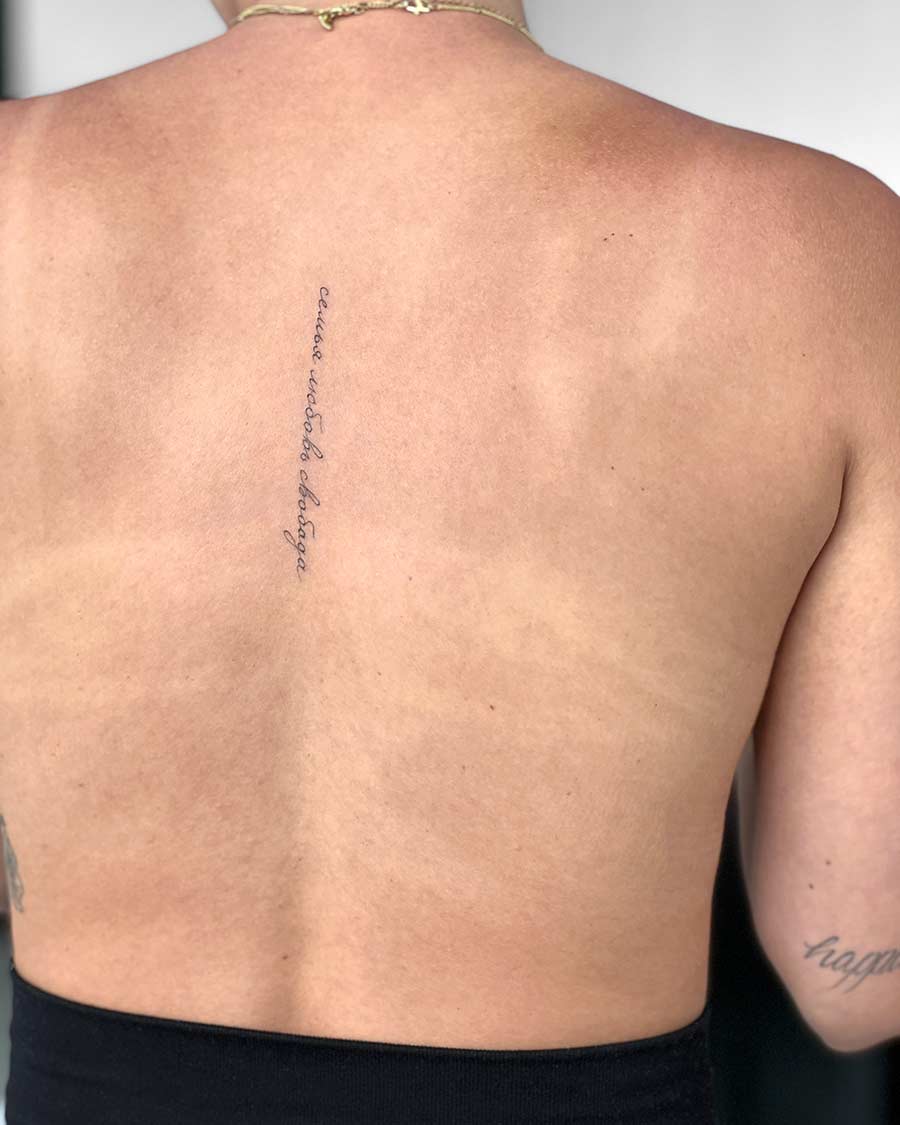 Close-up of a person's back with a vertical tattoo in cursive script reading "семья любовь свобода всегда" (family love freedom always) running along the spine.