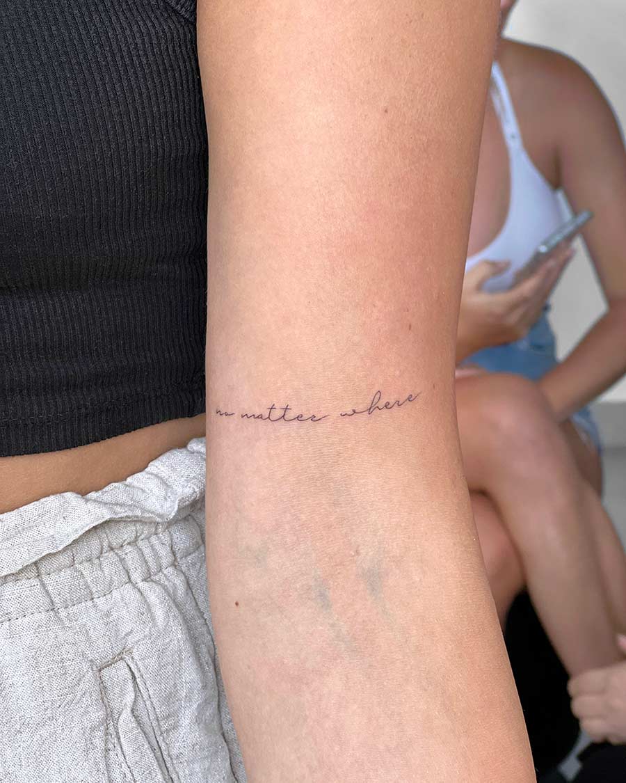 Close-up of a woman's arm with a delicate script tattoo reading "no matter where." Casual summer style with a black top and linen shorts visible.