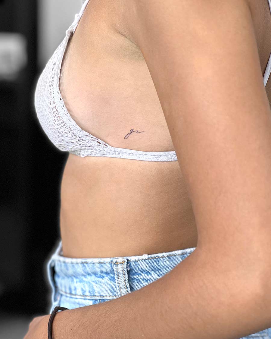 Close-up of a woman's torso wearing a white lace bralette and denim jeans, featuring a delicate "ji" tattoo on her ribcage. The tattoo is small and written in a cursive font.