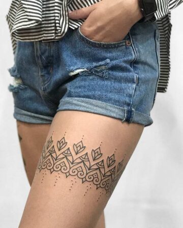 Close-up of a woman's leg with an ornamental tattoo design featuring geometric and floral patterns. She wears denim shorts and a striped top, hand in pocket.
