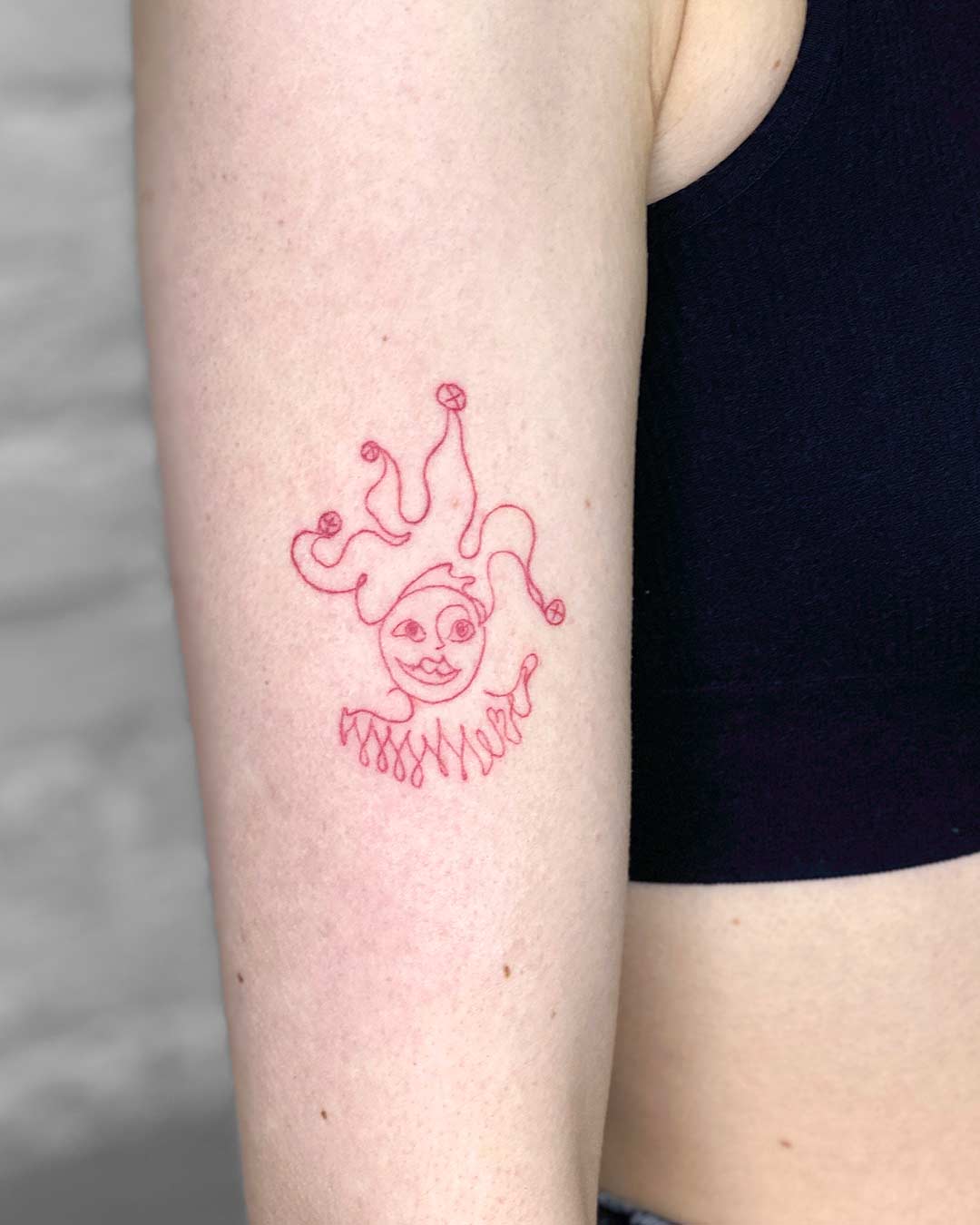 A minimalist red jester tattoo on an arm. The whimsical design features a smiling face, a ruffled collar, and a playful hat. This unique jester tattoo is a striking example of minimalist tattoo art. Done by tattoo artists in our fine line tattoo studio parlour shop Canggu, Bali
