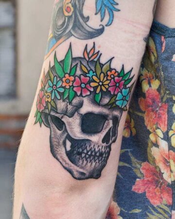Skull tattoo with a colorful flower crown on an arm. The tattoo features detailed shading and is surrounded by other tattoos and a floral shirt.