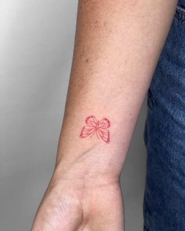 A small, delicate red butterfly tattoo on a woman's wrist. The minimalist design features fine lines, showcasing the butterfly's wings against the skin. She wears blue denim jeans. Done by tattoo artists in our fine line tattoo studio parlour shop Canggu, Bali