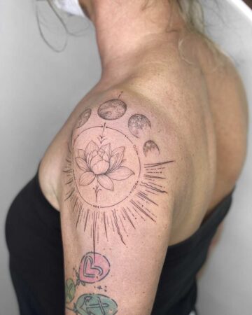 Shoulder tattoo featuring a delicate lotus flower surrounded by a circular design with moon phases and radiating lines, on a woman