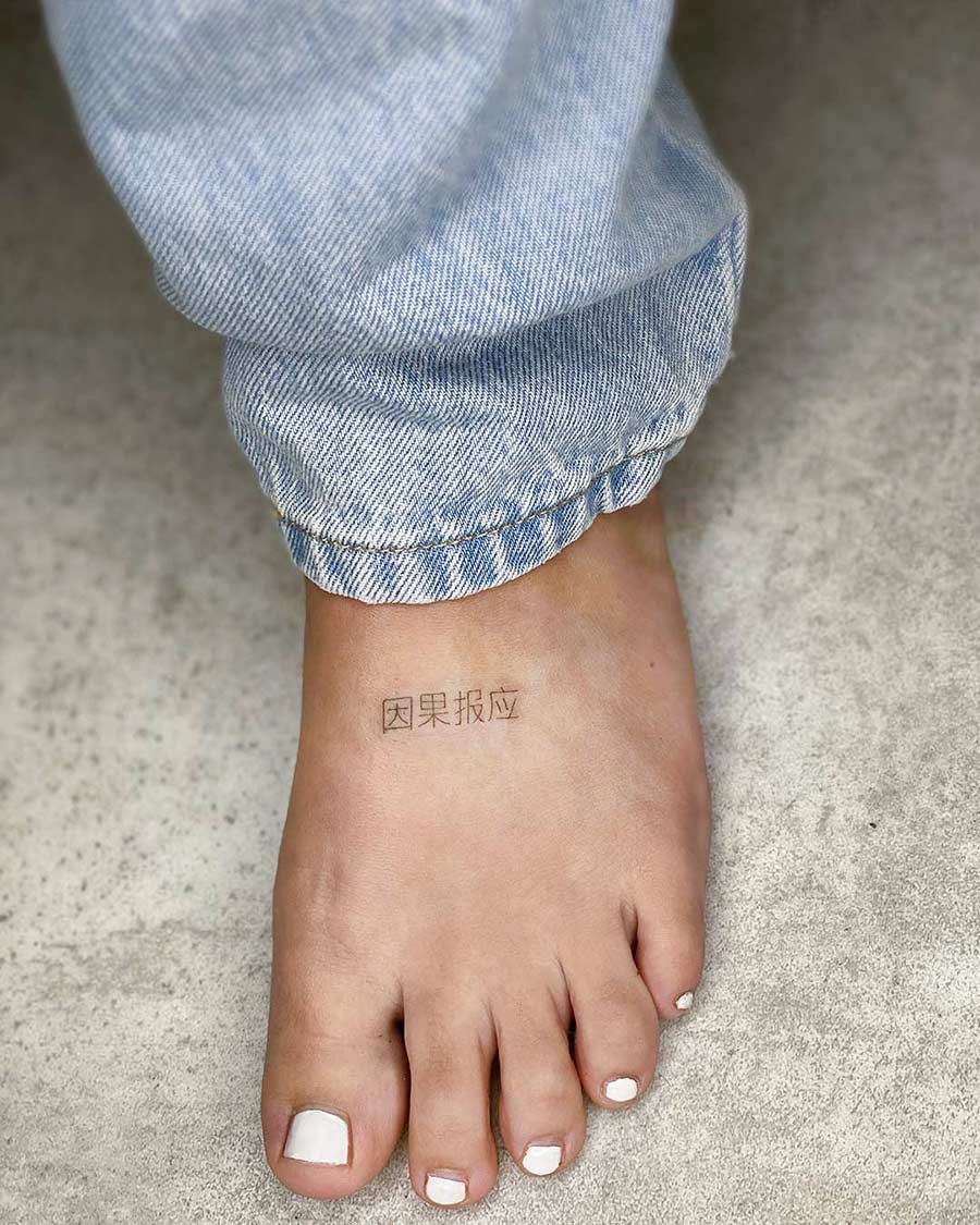 Foot with white pedicure and jeans. Tattoo on top of foot reads "因果报应", representing the concept of karma.