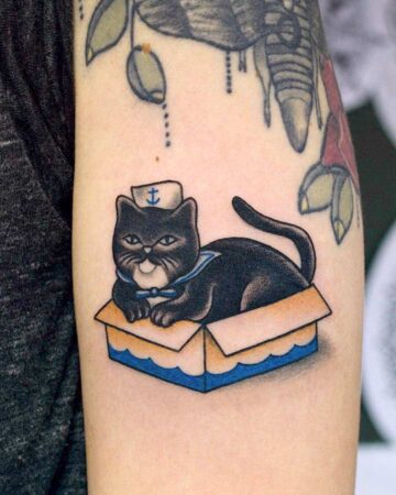 Tattoo of a black cat wearing a sailor hat and collar, sitting in a cardboard box with blue trim. Other nautical tattoos are visible in the background.