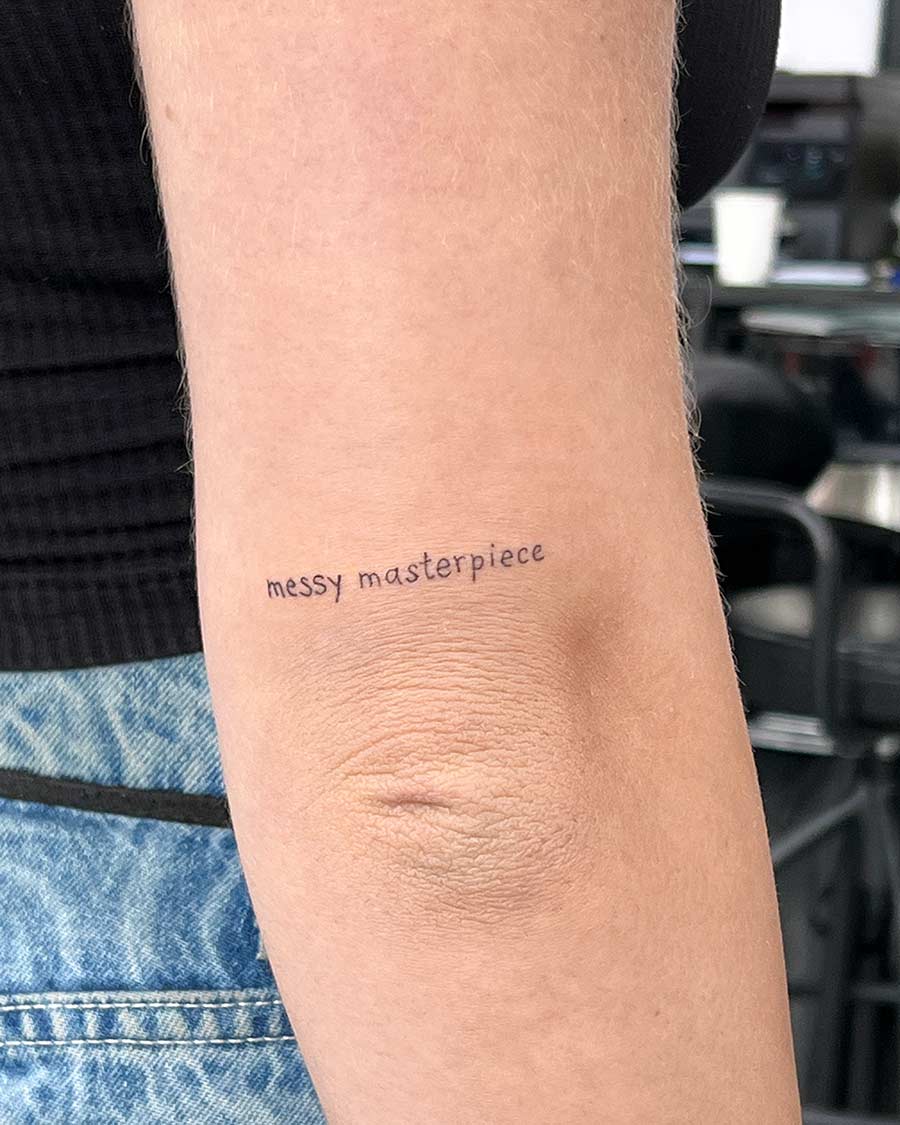 Close-up of an arm with a delicate "messy masterpiece" tattoo just above the elbow. The skin is fair, and the tattoo is in a fine, handwritten-style font. The arm is set against a blurred background.