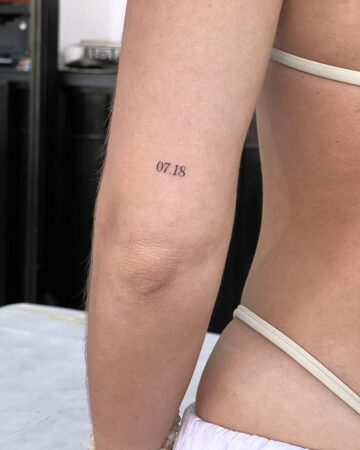 Close-up of a woman's arm and shoulder, featuring a small, minimalist tattoo of the date "07.18" in a simple sans-serif font, positioned above the elbow.