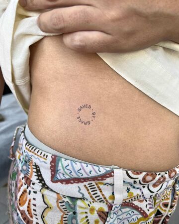 Close-up of a delicate tattoo on a person's side, reading "Saved By Grace" in a circular design. The individual is wearing patterned pants and a white top.
