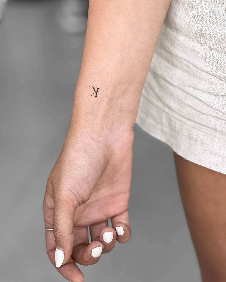 Close-up of a woman's hand and wrist featuring a delicate, minimalist tattoo of the letter "K" followed by a period. Her nails are painted white, and she's wearing a light-colored linen dress.