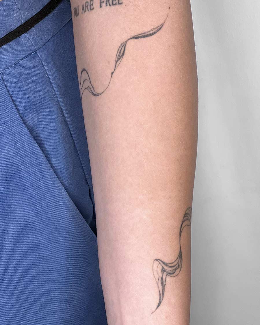 Close-up of a forearm with two minimalist, fine line tattoos. One tattoo curves along the arm, resembling flowing ribbons or smoke. The other is positioned further down the arm. The words "YOU ARE FREE" are partially visible above the upper tattoo. Blue fabric is visible on the left side of the image. Done by tattoo artists in our fine line tattoo studio parlour shop Canggu, Bali