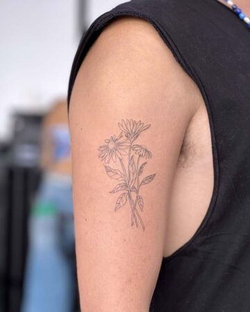 Close-up of a minimalist flower tattoo on a person's upper arm. The delicate line art depicts three daisies with leaves and stems, a subtle and elegant arm tattoo design. Done by tattoo artists in our fine line tattoo studio parlour shop Canggu, Bali