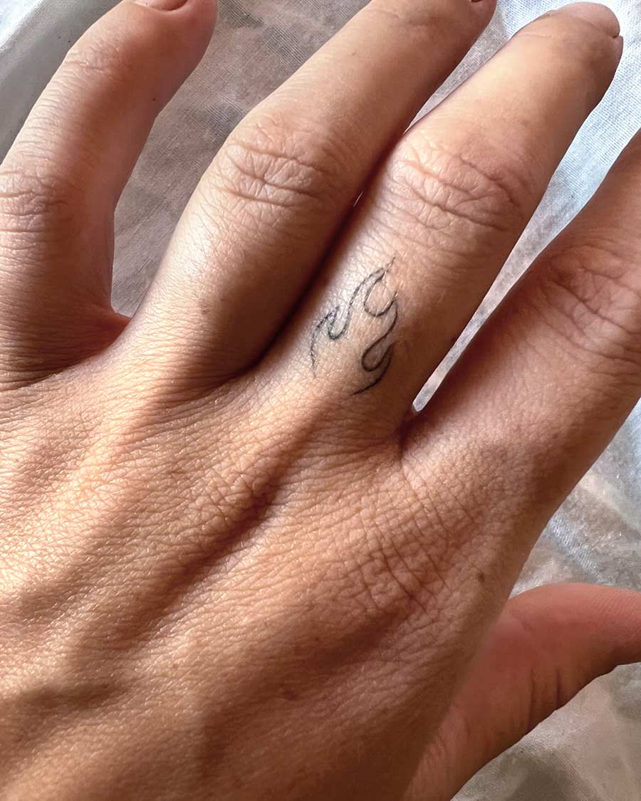 Close-up of a hand with a small flame tattoo on the ring finger. The minimalist tattoo features thin black lines, and the hand is relaxed, showcasing skin texture and detail. Done by tattoo artists in our fine line tattoo studio parlour shop Canggu, Bali