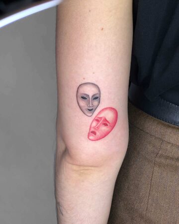 A tattoo featuring comedy and tragedy masks, one gray and smiling, the other red and sad. This body art expresses the duality of human emotion, a common theme in tattoo designs. Done by tattoo artists in our fine line tattoo studio parlour shop Canggu, Bali