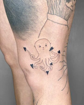 A cute octopus tattoo with heart-shaped suckers on a person's knee. The black ink design features a smiling octopus with curled tentacles, showcasing body art and a unique heart tattoo. Done by tattoo artists in our fine line tattoo studio parlour shop Canggu, Bali