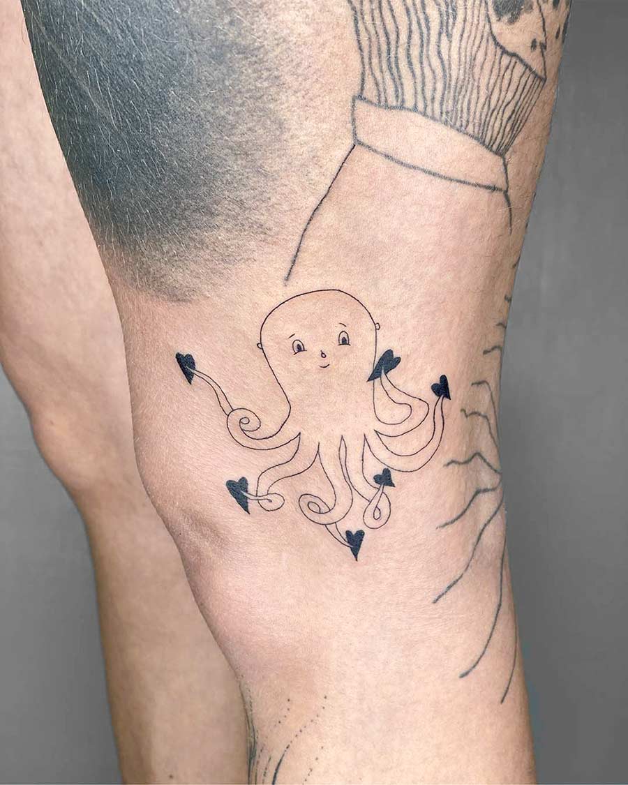 A cute octopus tattoo with heart-shaped suckers on a person's knee. The black ink design features a smiling octopus with curled tentacles, showcasing body art and a unique heart tattoo. Done by tattoo artists in our fine line tattoo studio parlour shop Canggu, Bali