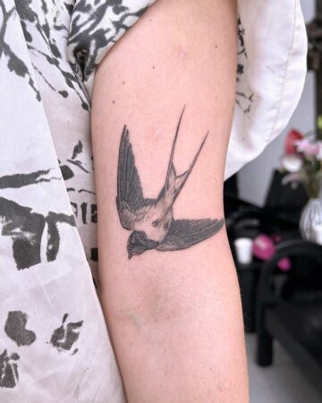 Detailed black and grey swallow tattoo on a person's arm. The bird is depicted in flight, wings spread wide, showcasing intricate feather details. A popular swallow tattoo design. Done by tattoo artists in our fine line tattoo studio parlour shop Canggu, Bali