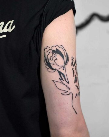 A striking minimalist tattoo of a stylized rose on an arm. The flower's petals are filled with solid black, while the stem and leaves are rendered in delicate line art. A line art tattoo design featuring tiny figures adorns the stem, adding a unique detail. Perfect example of a minimalist tattoo. Done by tattoo artists in our fine line tattoo studio parlour shop Canggu, Bali