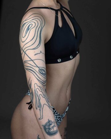 A woman with intricate arm tattoos, including abstract line work and whimsical figures, poses confidently in a black bikini top and leopard print bottoms. Her tattoos are a striking display of body art, celebrating body positivity and individual expression with ink. Done by tattoo artists in our fine line tattoo studio parlour shop Canggu, Bali