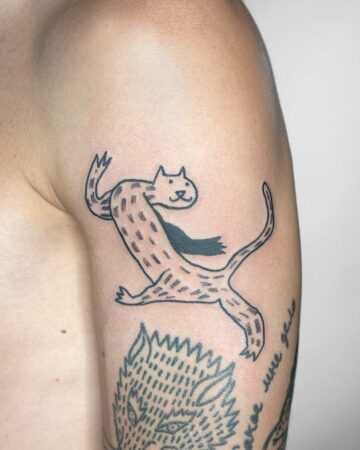 A whimsical cat tattoo in black ink on a person's arm. The minimalist design features a stylized cat in mid-stride, with a simple black scarf. Another tattoo is visible below the cat, adding to the unique tattoo design aesthetic. done by artists in our fine line tattoo studio Canguu Bali